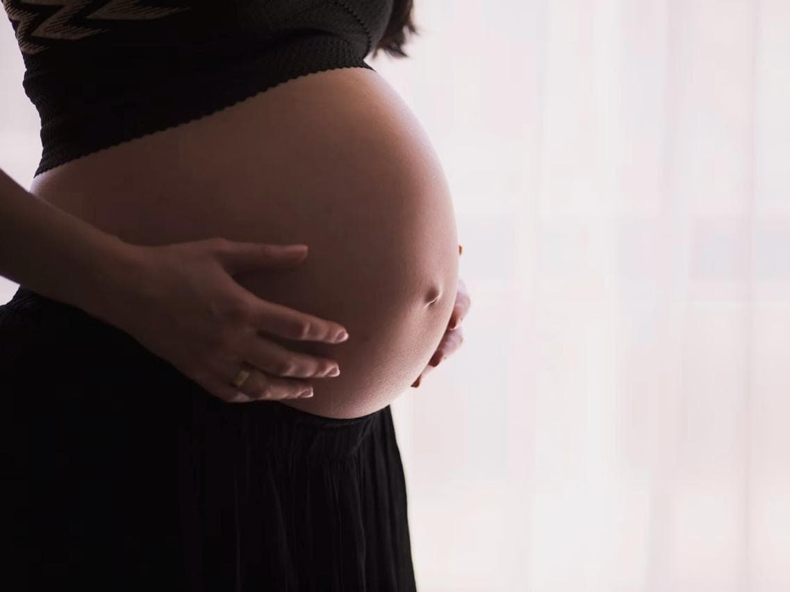 A pregnant woman standing by a bright window, both hands resting on her belly