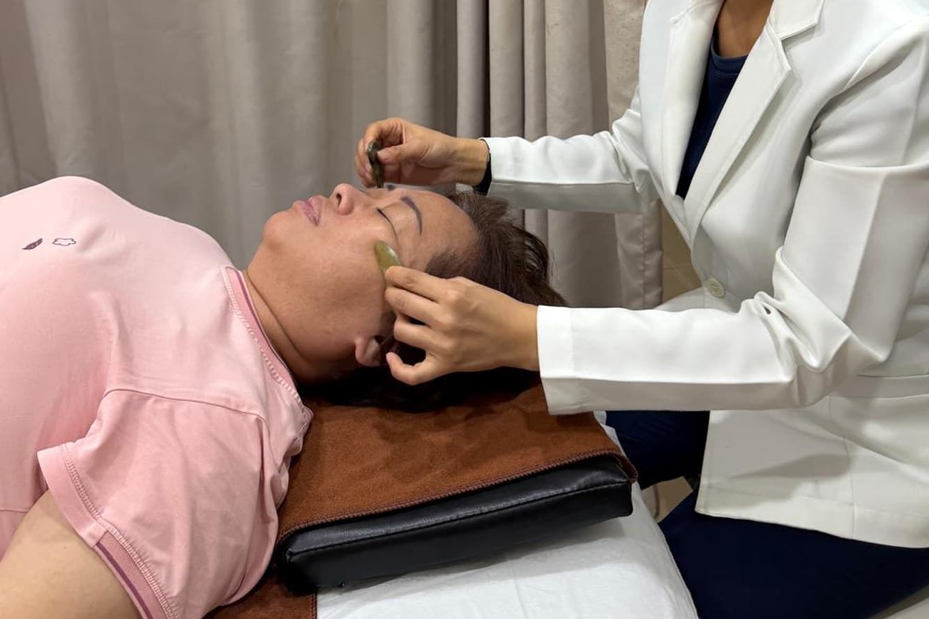 A physician drawing a jade gua sha tool along a patient's cheek, the patient lying on the couch with eyes closed.